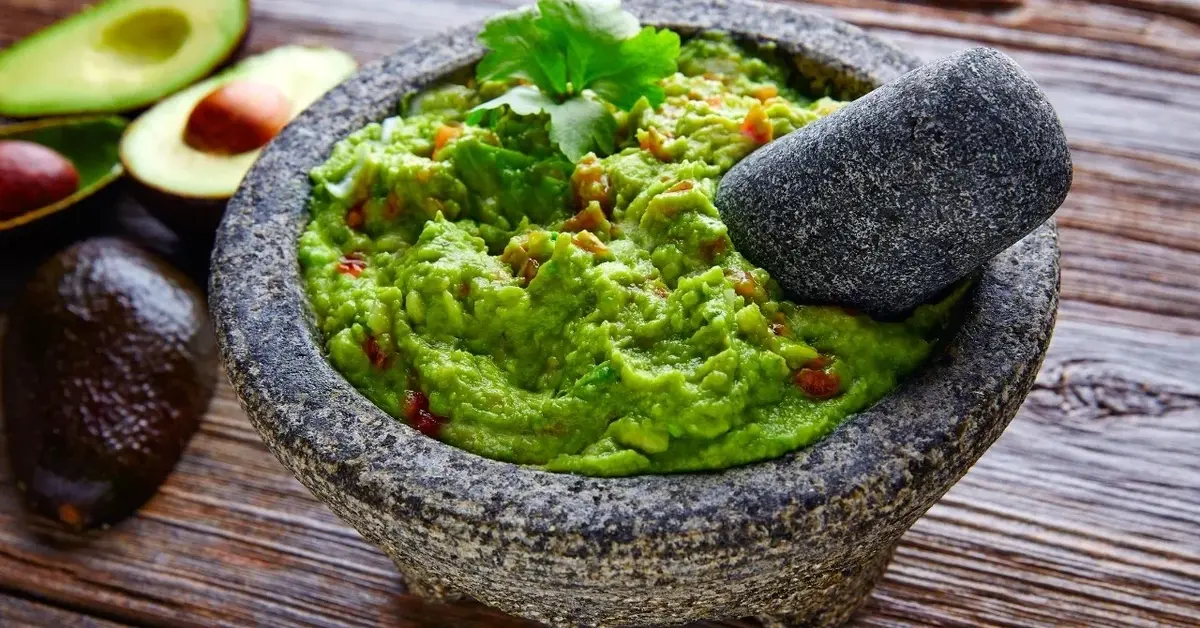 guacamole w moździerzu kamiennym na tle ciemnego drewna obok połówki awokado