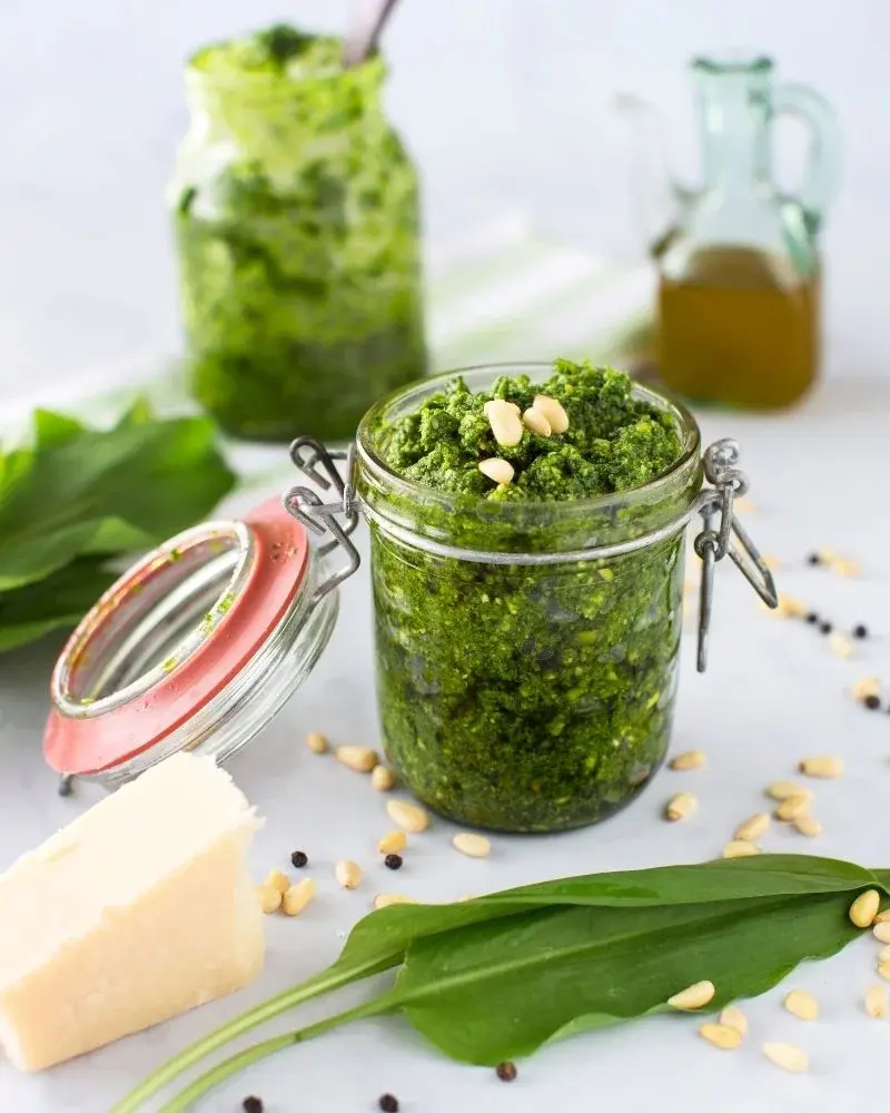 Pesto z czosnku niedźwiedziego w słoiku, obok rozsypane orzeszki, olej i kawałek sera dojrzewającego