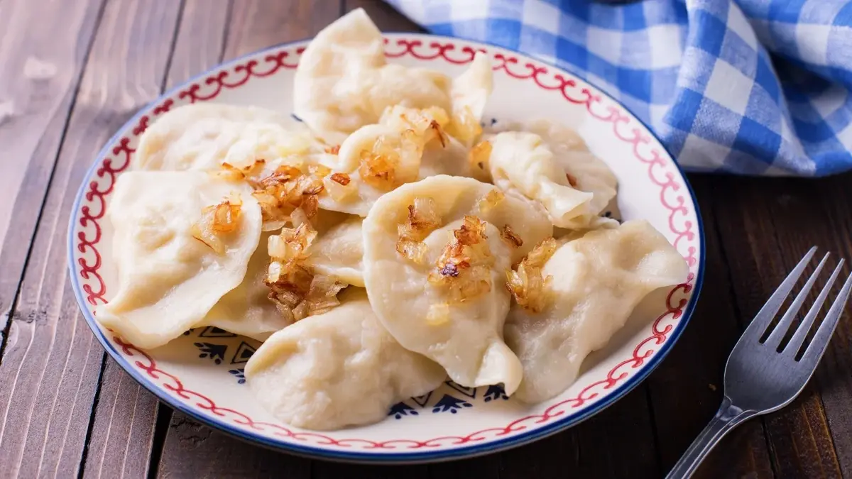 pierogi posypane cebulą w ozdobnym talerzu na drewnie obok ścierka w kratkę niebieska i widelec