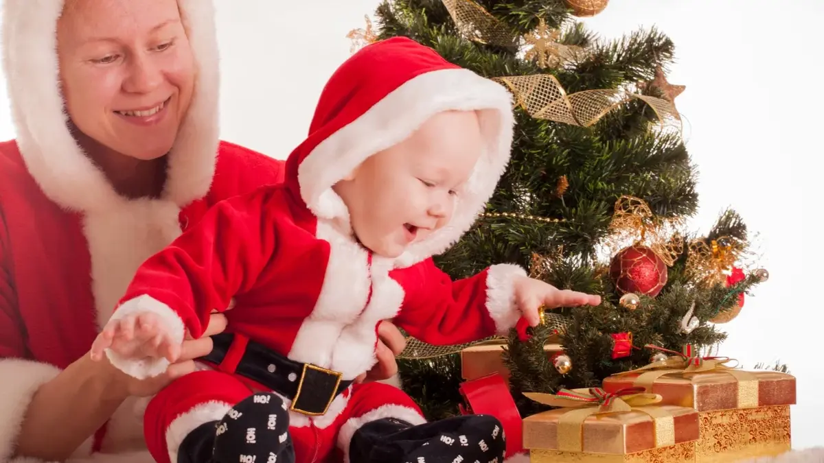 niemowlę sięga uśmiechnięte po prezent, trzyma go mama, z tyłu choinka