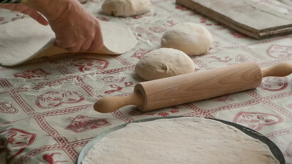 Ciasto na pizze rozwałkowane okrągłe. Wałek drewniany i ręce podnoszące rozwałkowany placek. Trzy kulki ciasta. 