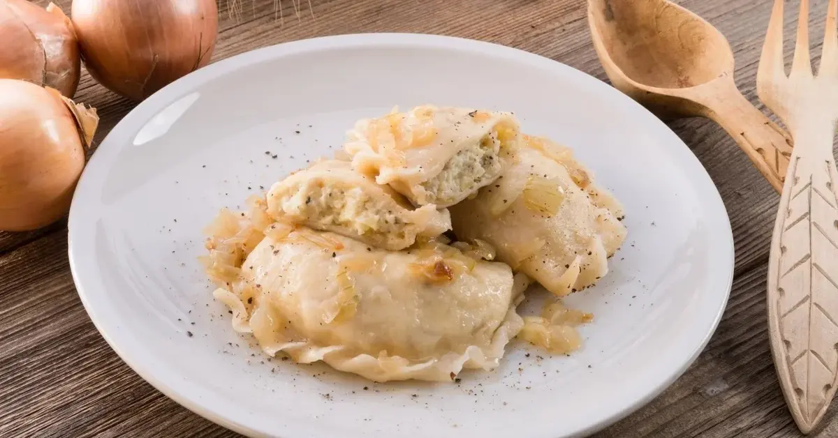 Talerz z trzema pierogami ruskimi, obok drewniany widelec i łyżka. Po drugiej stronie trzy cebule