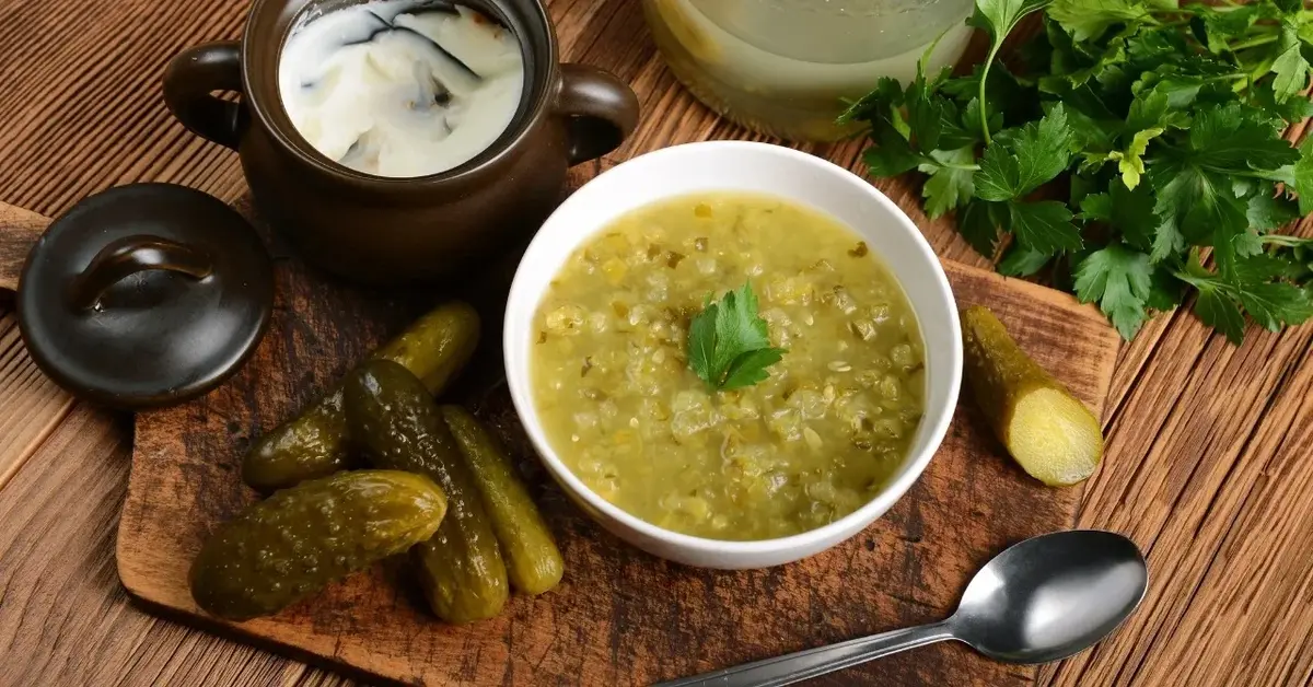 Zupa ogórkowa w białym głębokim talerzu, obok ogórki kiszone, pojemnik ze śmietaną i natka pietruszki. 