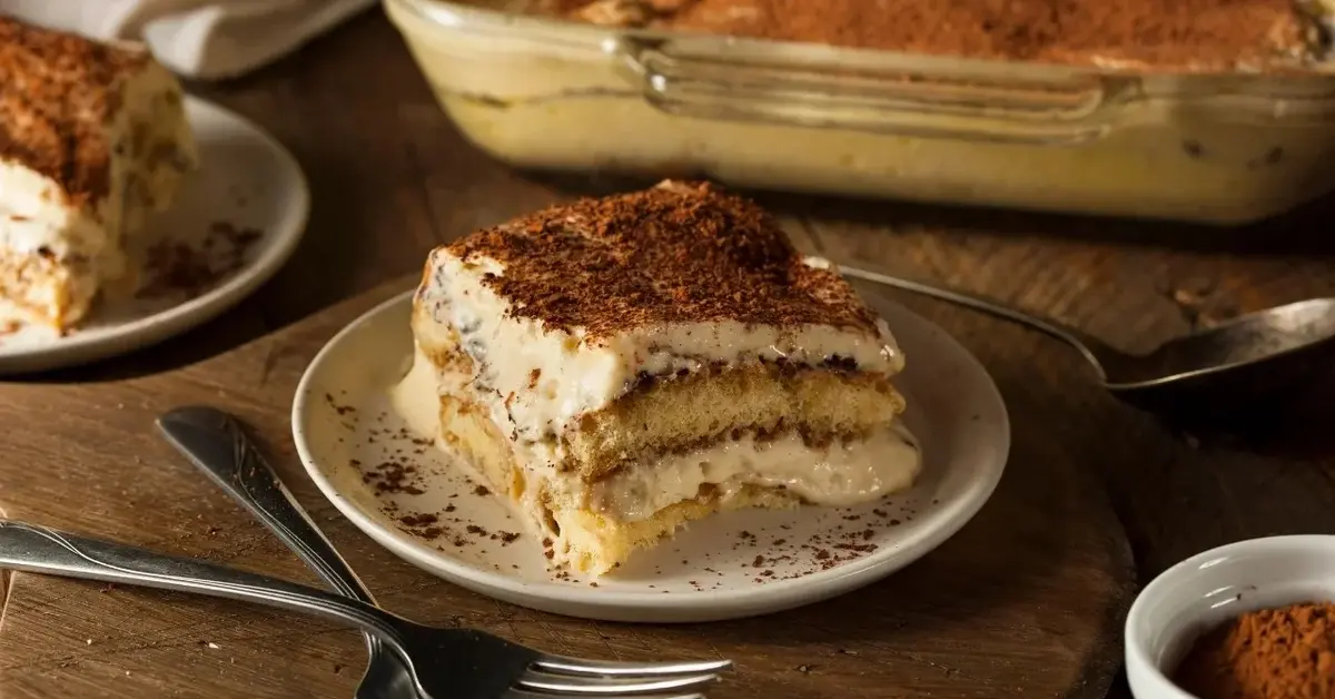 Tiramisu na małym talerzyku na drewnianym blacie. Obok srebrne sztućce, fragment większego pojemnika na tiramisu 