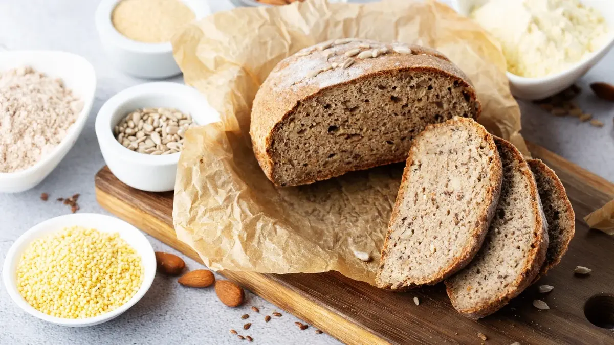 Bochenek bezglutenowego chleba i dwie kromki na desce drewnianej. Obok miseczki z różnymi mąkami