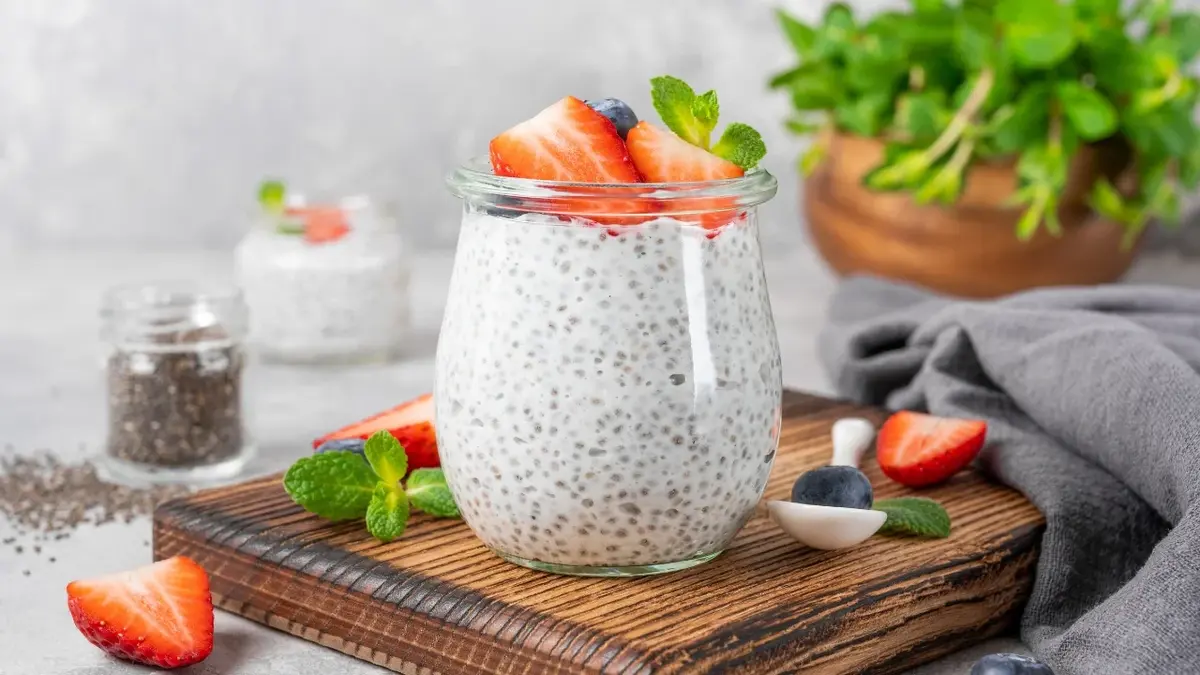 Chia pudding z owocami i orzechami w szklance na drewnianej desce. W tle truskawki, nasiona chia w słoiczku i zioła