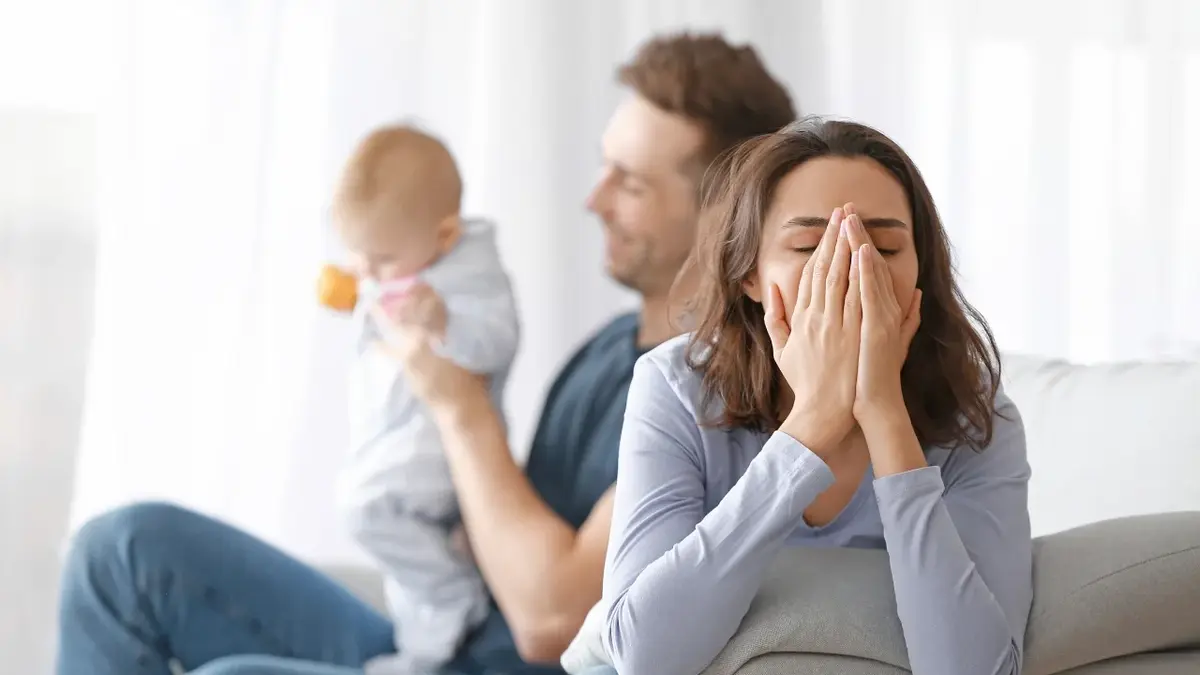 Kobieta zakrywająca twarz dłońmy, w tle mężczyzna trzymający na rekach niemowlę