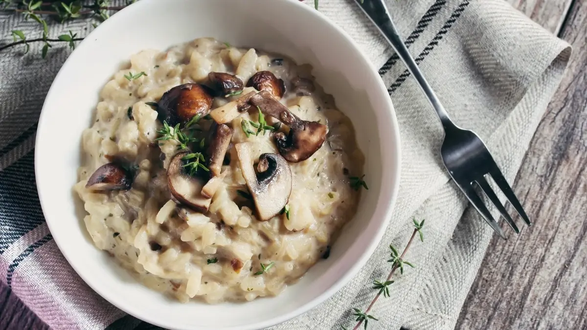 Risotto z pieczarkami w białej miseczce leżącej na drewnianym blacie. Obok biała ściereczka i widelec