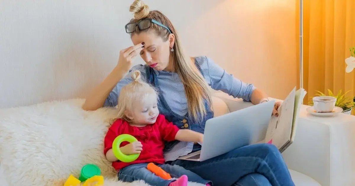 Mama zmęczona macierzyństwem siedzi z dzieckiem i pracuje.