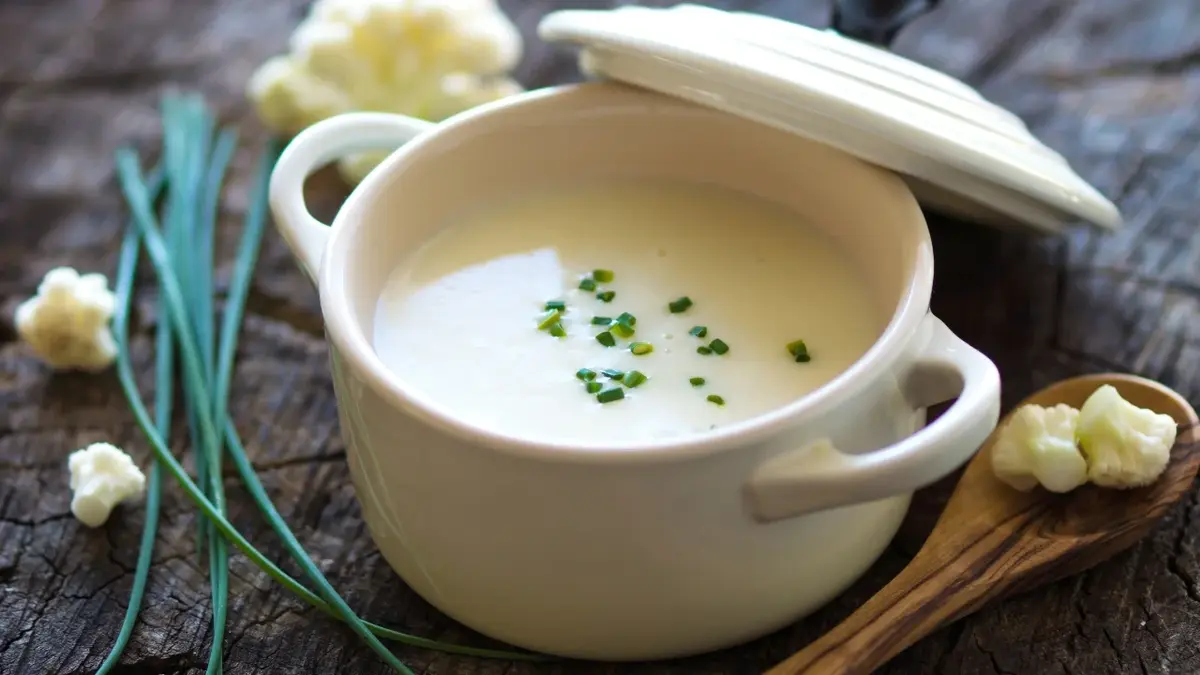 zupa kalafiorowa krem w garnku z pokrywką obok kawałki kalafiora szczypiorek i łyżka drewniana