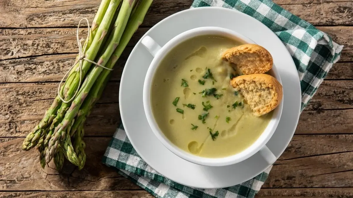 Zupa szparagowa. Obok szparagi. W talerzu z zupą jest pieczywo.