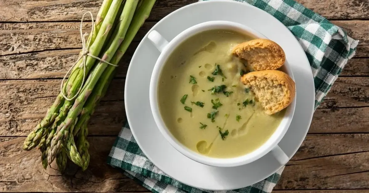 Zupa szparagowa. Obok szparagi. W talerzu z zupą jest pieczywo.