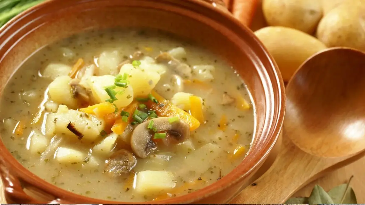 Zupa ziemniaczana z leśnymi grzybami podana w brązowej, rustykalnej misce, obok leży drewniana łyżka